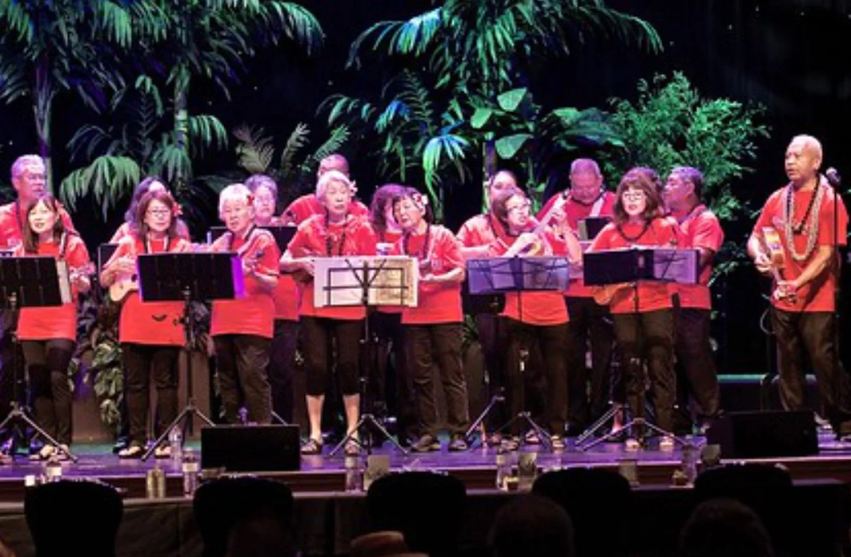 A group of people in red shirts playing ukuleles on stage with a tropical plant background.