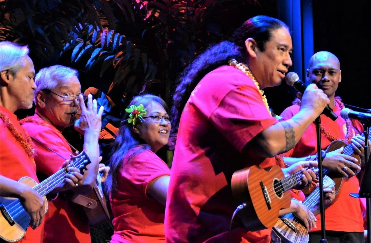 A group of musicians in red shirts playing ukuleles on stage with microphones.