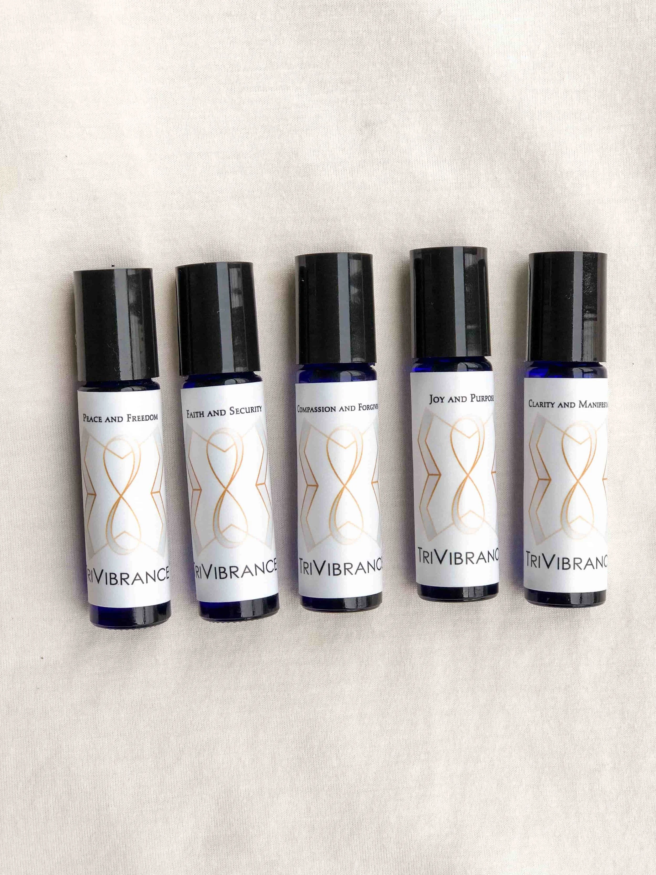Five essential oil roller bottles with labels reading Peace and Freedom, Faith and Security, Compassion and Forgiveness, Joy and Purpose, and Clarity and Manifestation, on a light-colored fabric background.
