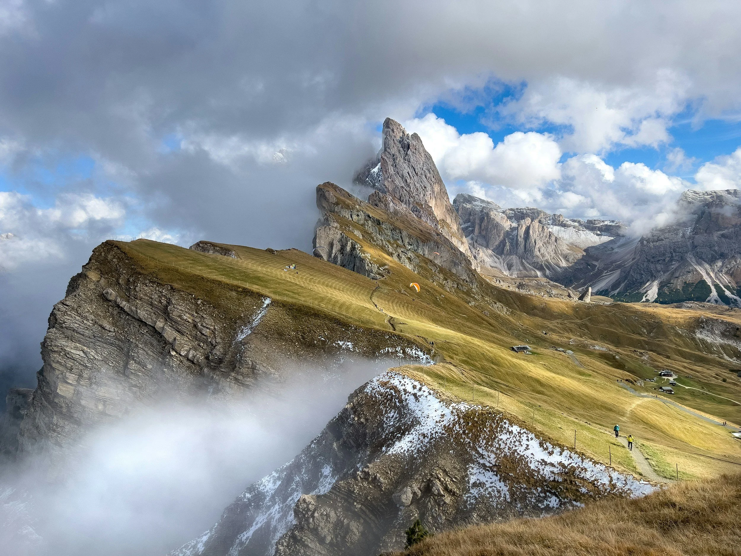 Scenic view of a mountain landscape with a grassy sloped ridge, rocky cliffs, and clouds surrounding the peaks. Several hikers are walking along a trail, and small buildings and a parachute or paraglider are visible in the distance.