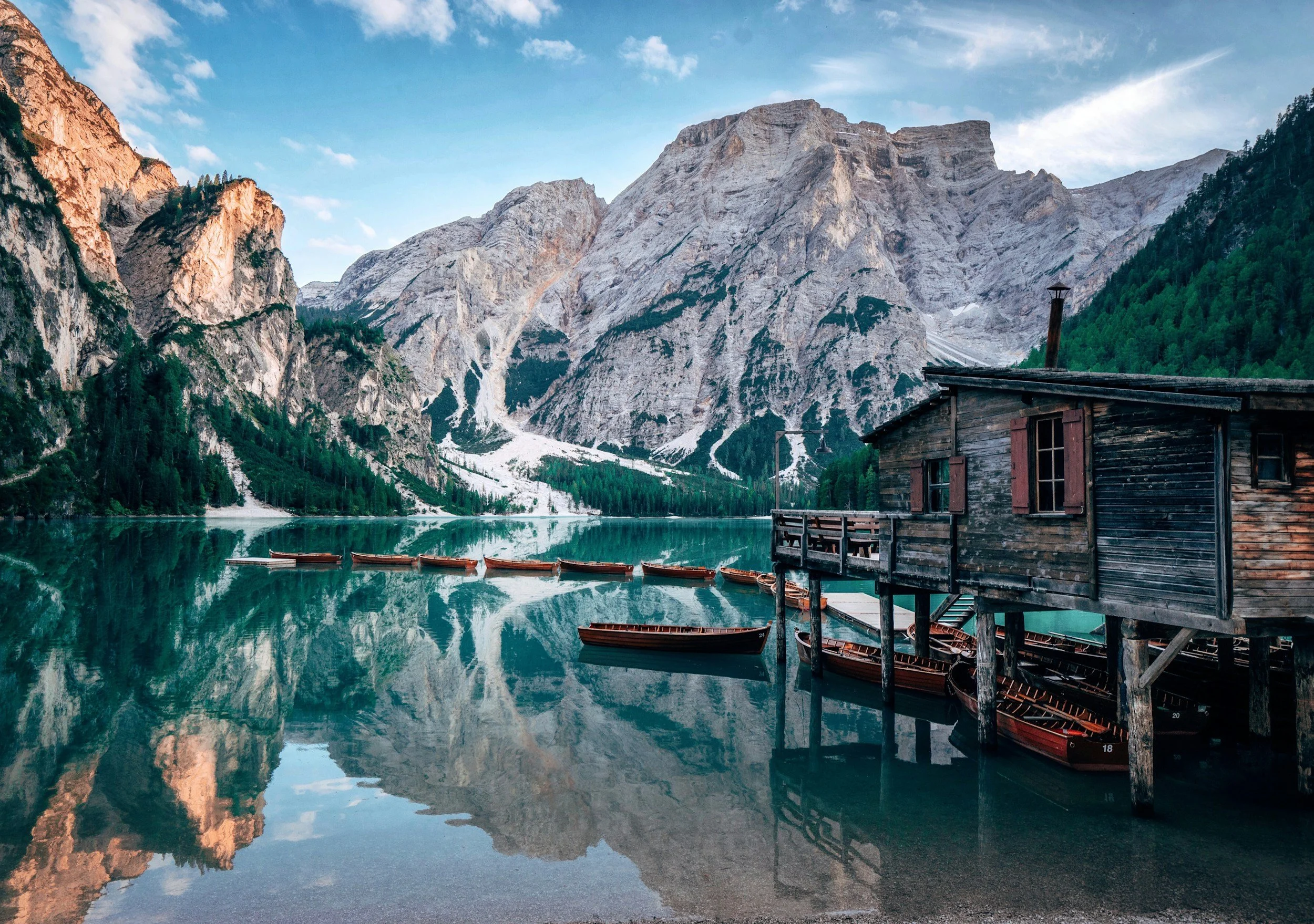 A rustic wooden boathouse on stilts extends over a serene lake, surrounded by towering mountains and dense green pine trees, with multiple wooden boats floating near the boathouse and a clear, partly cloudy sky overhead.