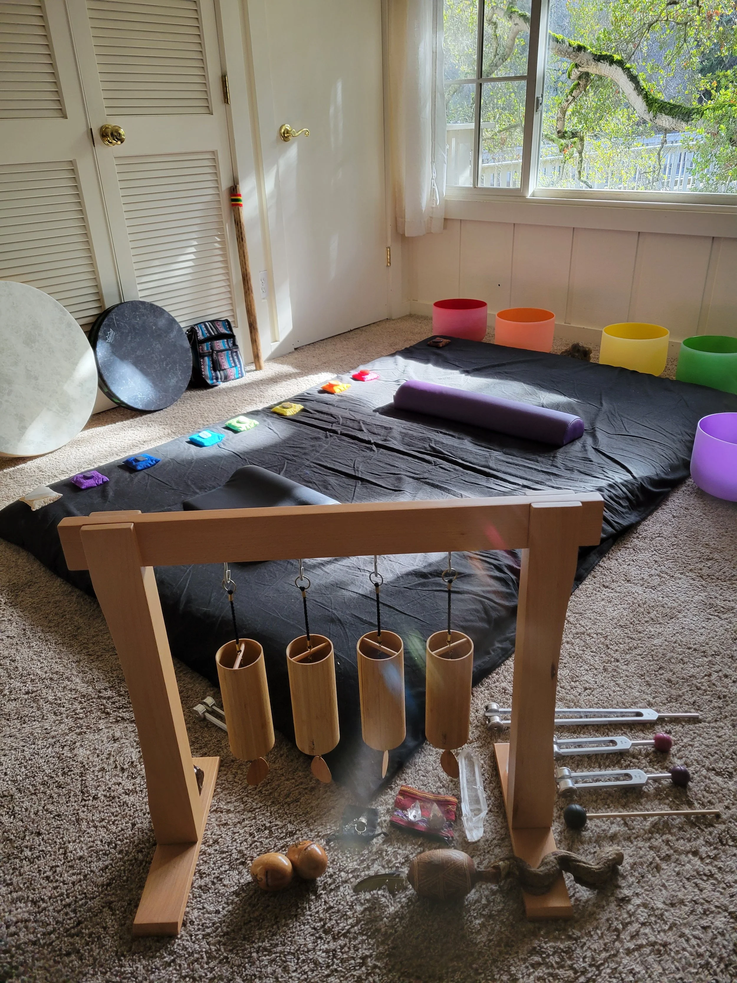 A room with a black mat on the carpeted floor, set up with colorful singing bowls, percussion instruments, and percussion mallets. There are wooden wind chimes hanging from a wooden stand in the foreground, and a view of trees through a large window.