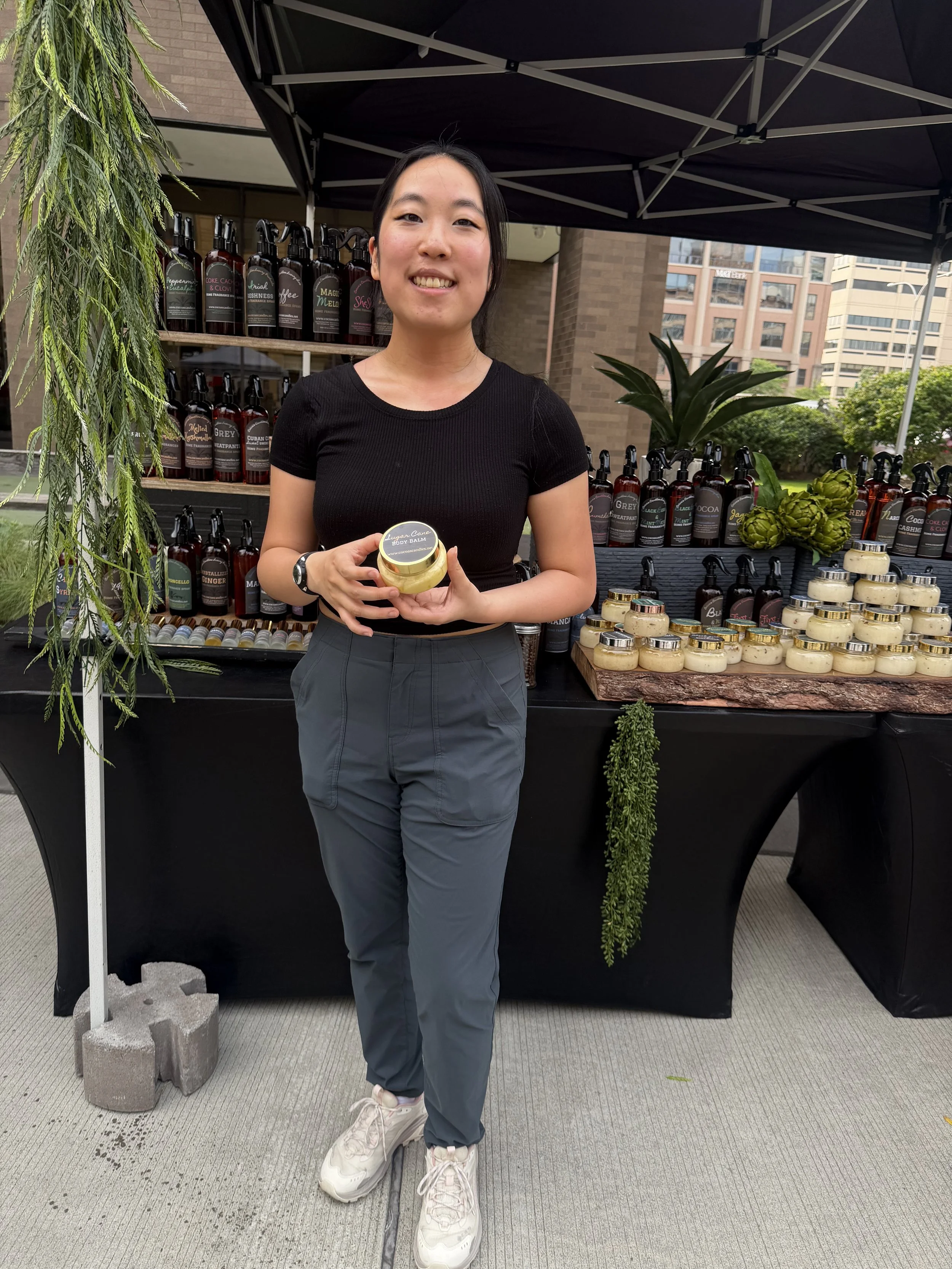 A young woman standing at an outdoor market stall with bottles of products and jars, holding a small jar and smiling at the camera.
