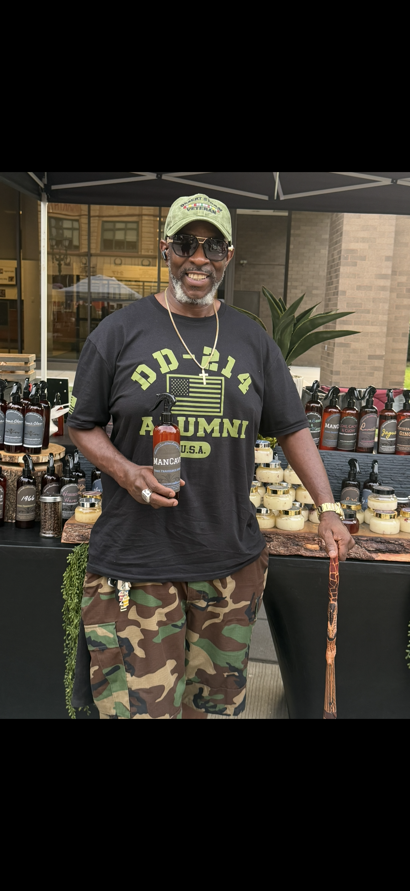 A man wearing a black T-shirt with military camouflage pattern shorts, sunglasses, and a green cap, stands outdoors behind a table displaying candles and lotion bottles. He is holding a bottle of fragrance oil and smiling.