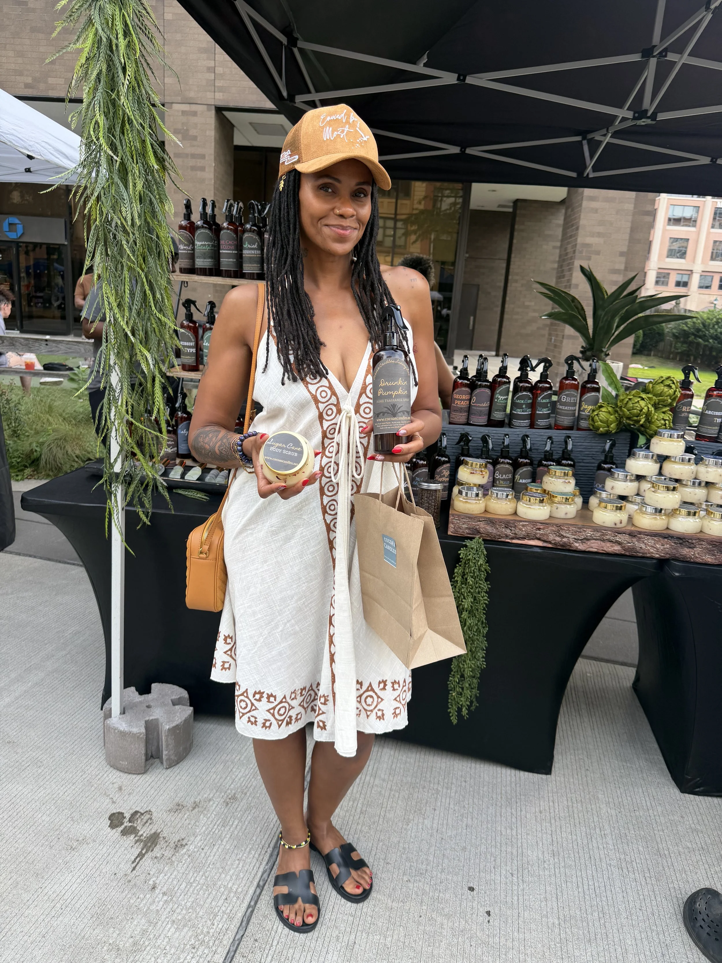 A woman stands outdoors at a market stall with beauty products. She wears a beige cap, a white dress with brown patterns, black sandals, and has a tattoo and red nail polish. She holds a body scrub container and a brown paper bag, smiling at the came
