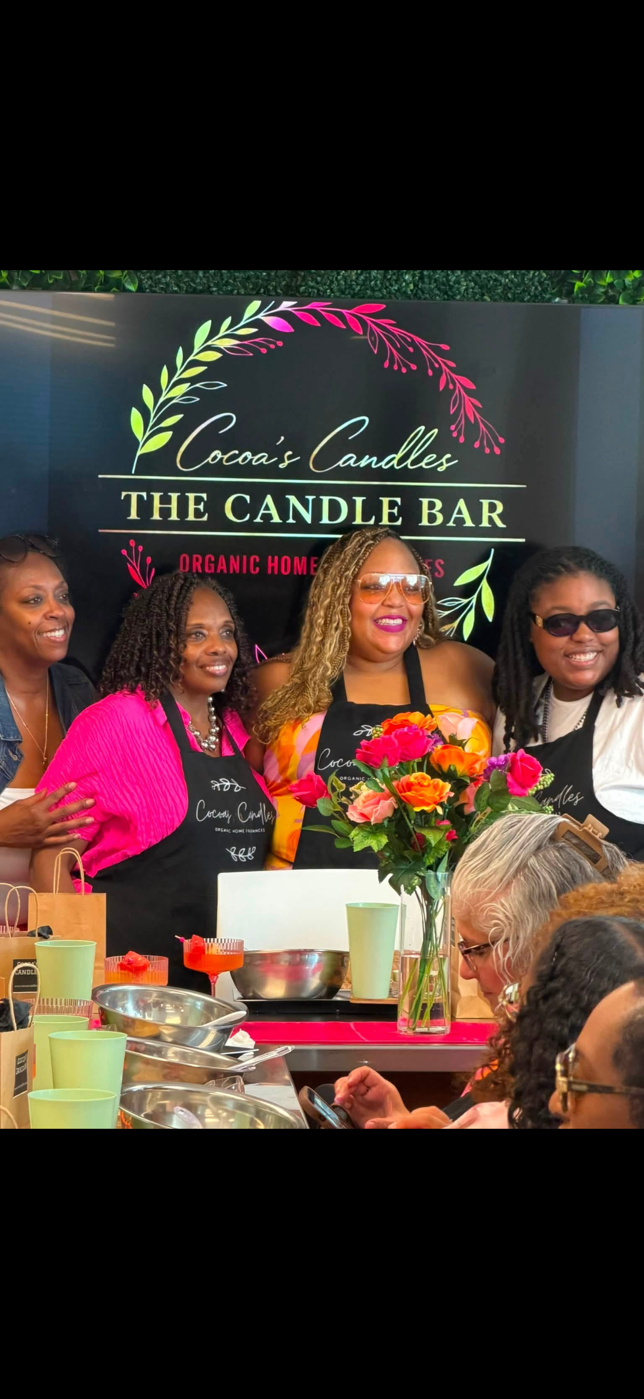 Group of women smiling at a cooking demonstration or class at Cocoa's Candles, a candle bar and organic home fragrances business, with floral decor and various kitchen utensils and ingredients on the table.