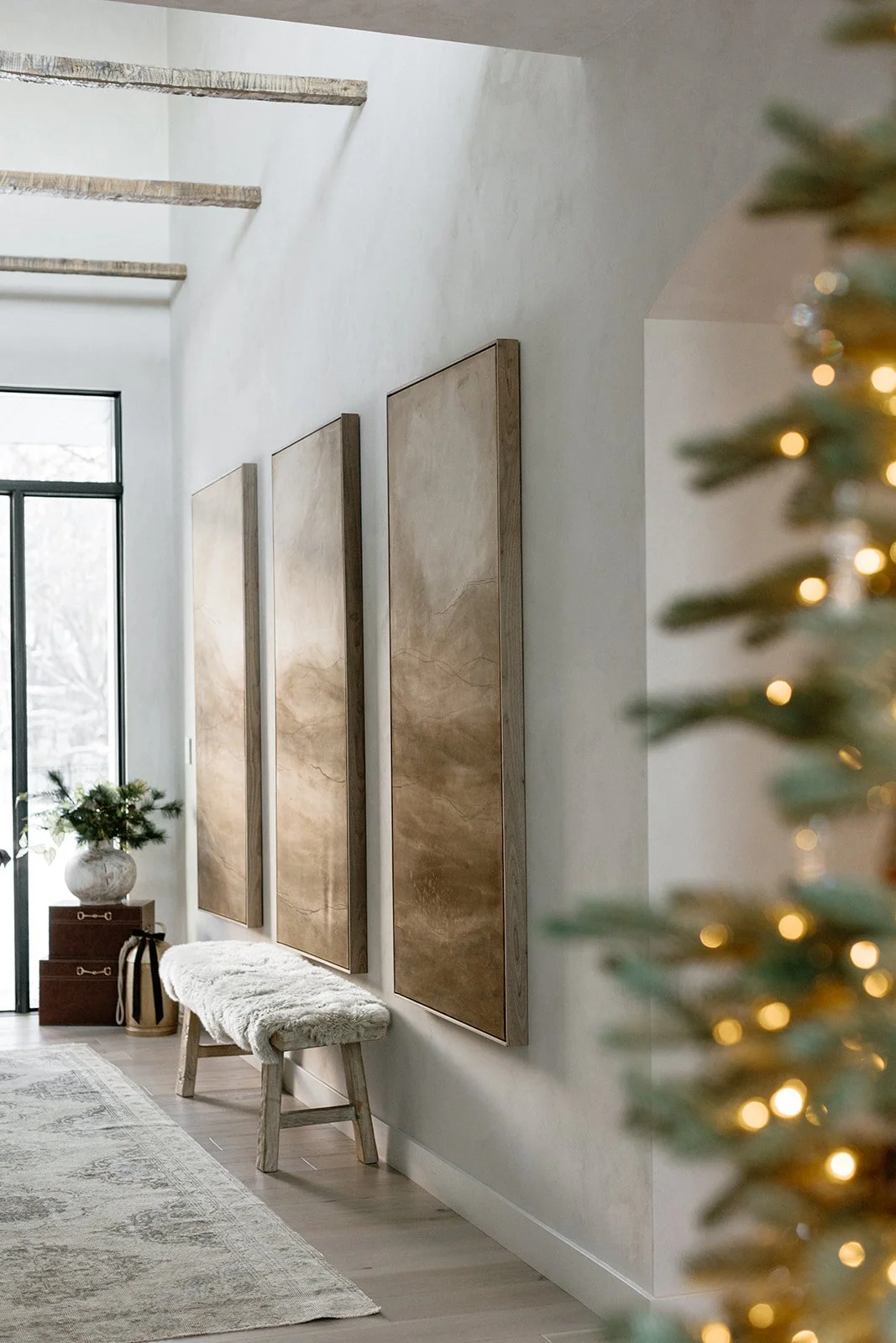 A modern, minimalistic interior space with three large, rectangular beige artwork pieces on a white wall. There is a white, fluffy bench below the artworks, a small side table with decorative items, and a Christmas tree with lights at the right edge 