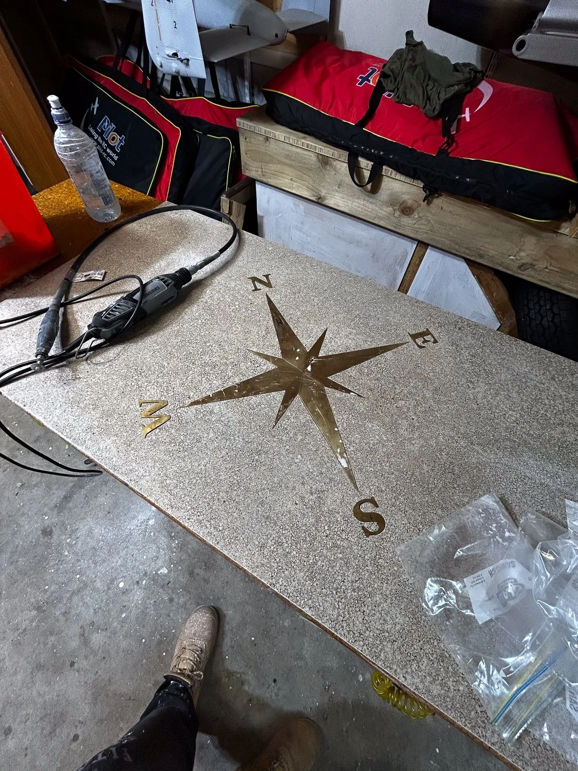 A workbench with a metal compass rose design and letter markings for directions. There is a water bottle, a grinder tool, and a plastic bag with parts on the bench. In the background, there are sports bags, a bed, and some clothing.