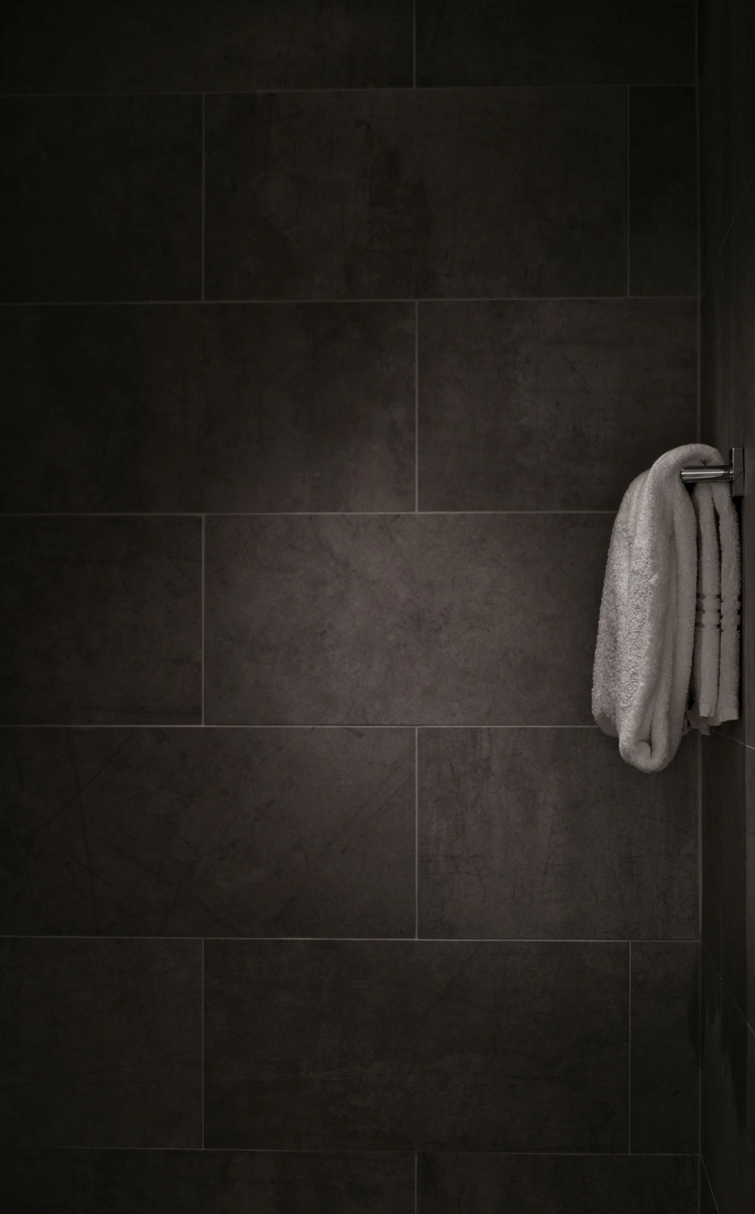 Dark tiled wall with a folded gray towel hanging on a silver towel rack on the right side.