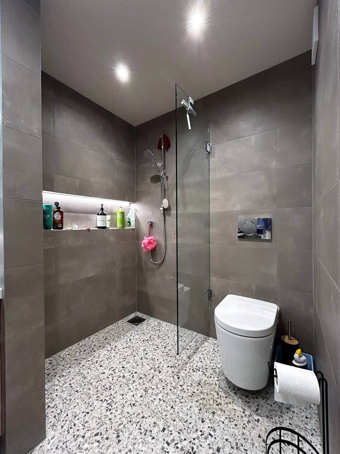 Modern bathroom with gray tiled walls and terrazzo floor, featuring a walk-in shower with a glass partition, a wall-mounted toilet, and a built-in shelf with toiletries.