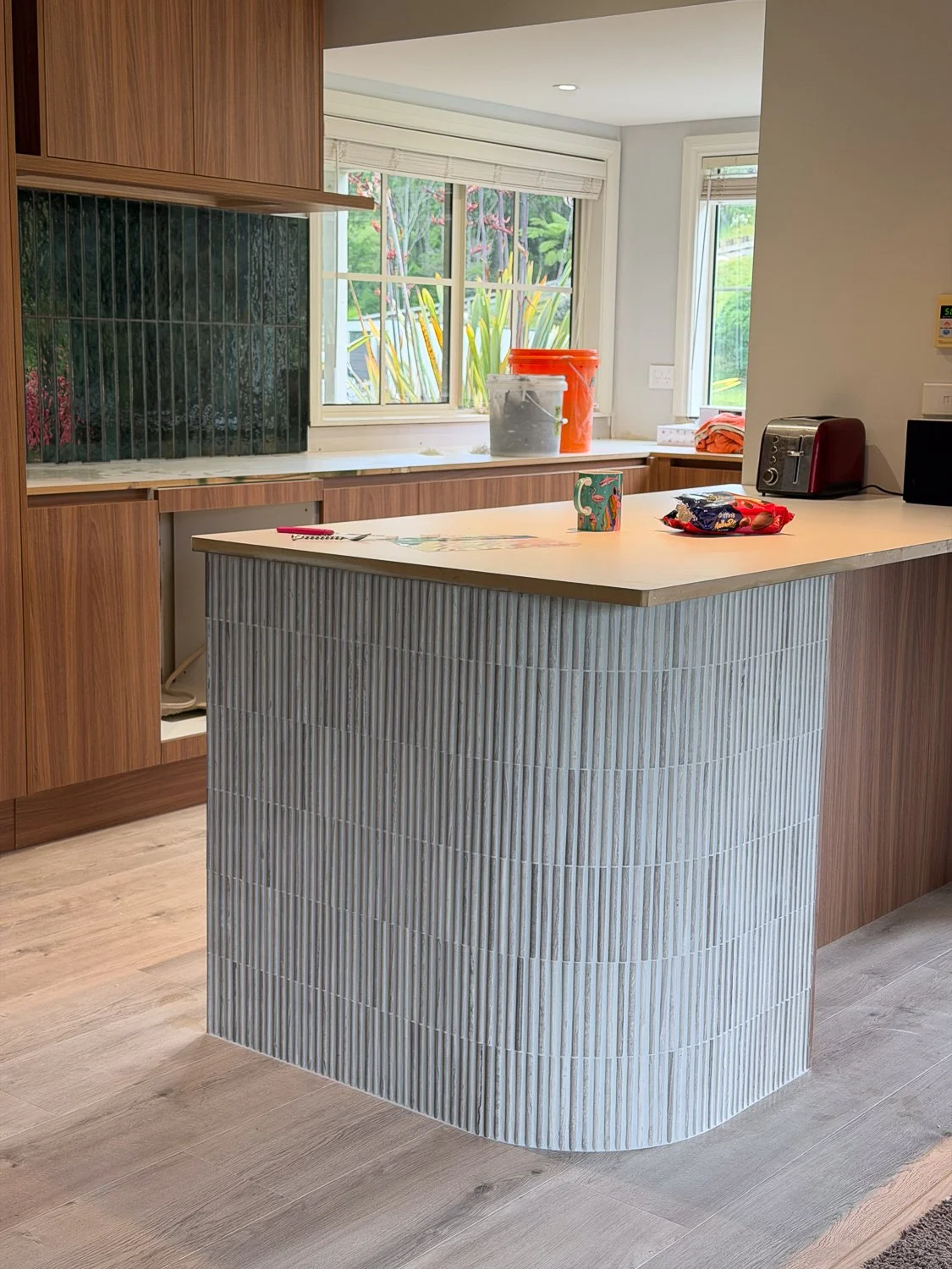 A kitchen with a curved counter covered in small light-colored tiles, wooden cabinets, and a window showing outdoor greenery.