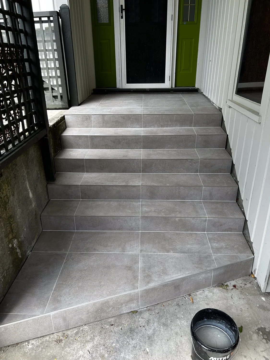 Gray tiled stairs leading up to a door with a green frame, side wall with vertical white siding, and black metal fencing on the left. A black bucket with gray water on the concrete ground in front.