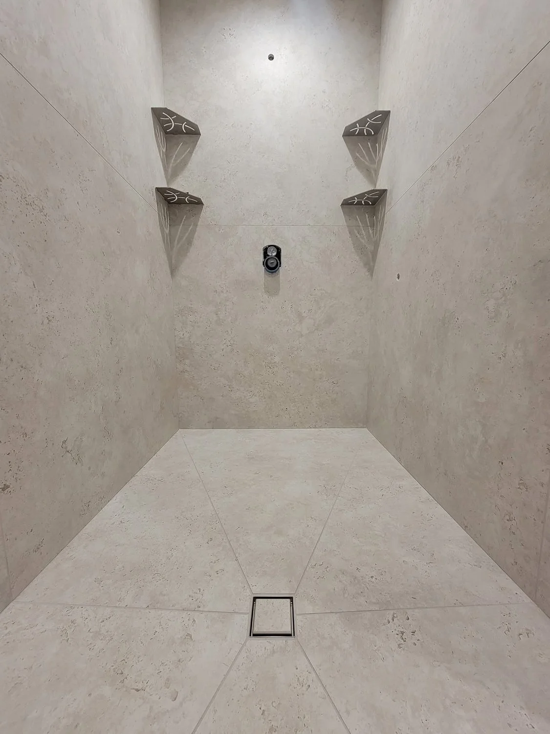 Empty shower with beige tiled walls and floor, two shelf brackets on the left wall, a showerhead missing on the right wall, and a drain in the floor.