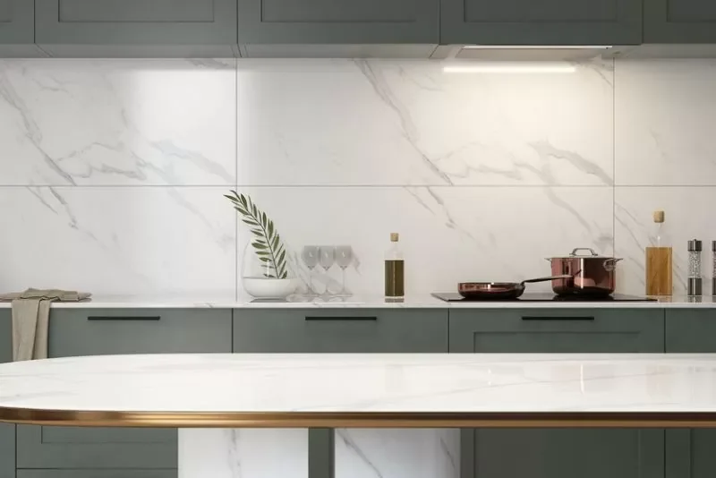 Modern kitchen with gray cabinets, white marble backsplash, and a white marble countertop with a gold edge. Decor includes a potted plant, wine glasses, a bottle of olive oil, a pink pot on the stove, and salt and pepper shakers.
