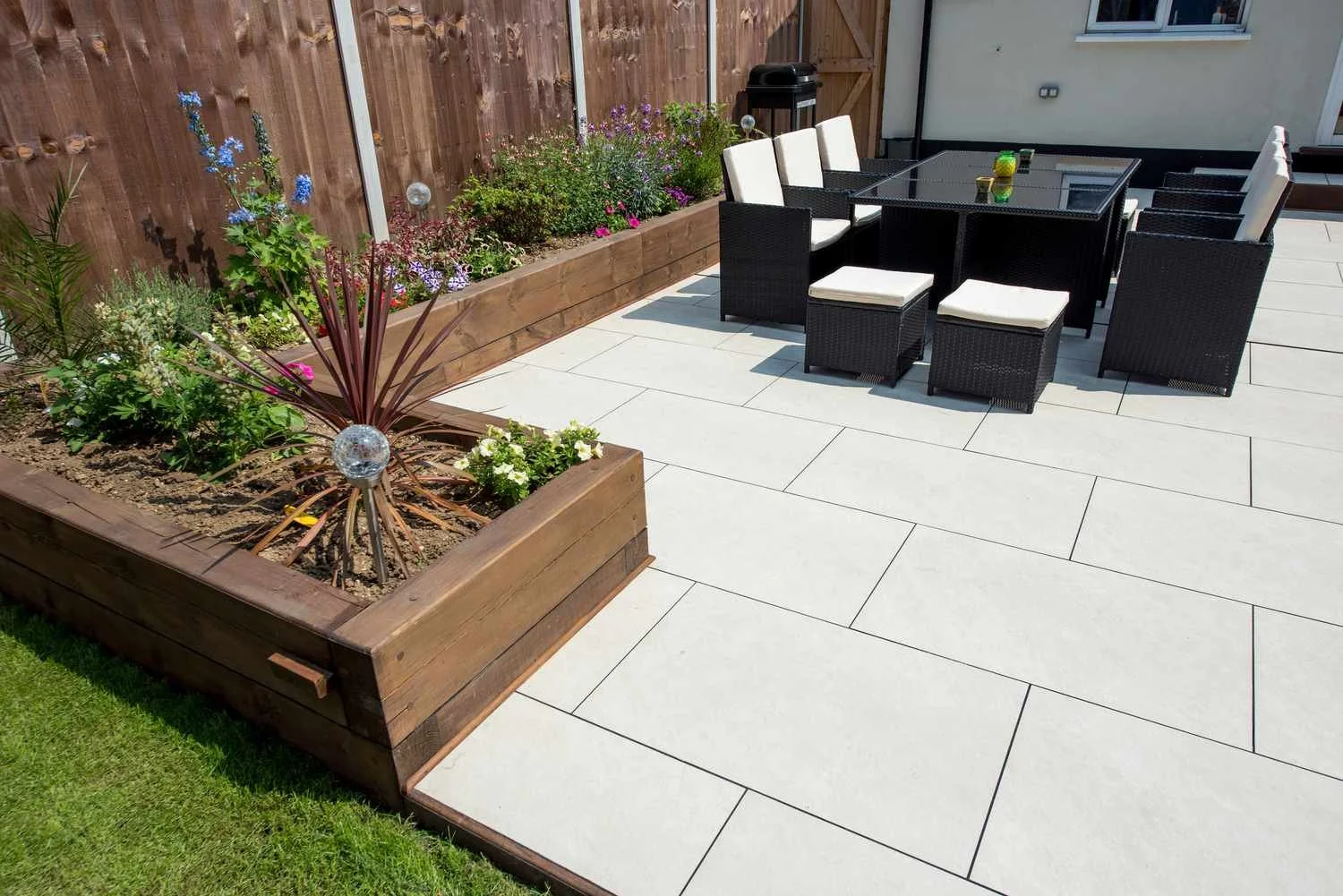 Back patio area with a wooden fence, a flower bed with various plants, a black outdoor dining table with six chairs and two ottomans, a barbecue grill, and a window on a house wall.
