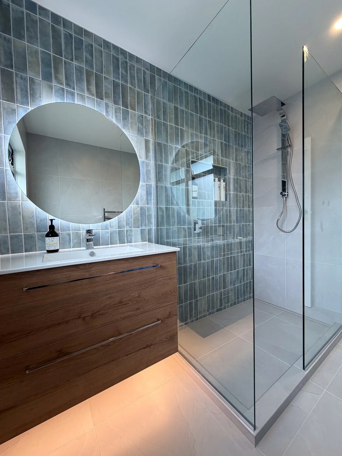 Modern bathroom with a glass-enclosed shower, wooden vanity with a round mirror, gray tiled walls, and a bottle of soap on the sink.