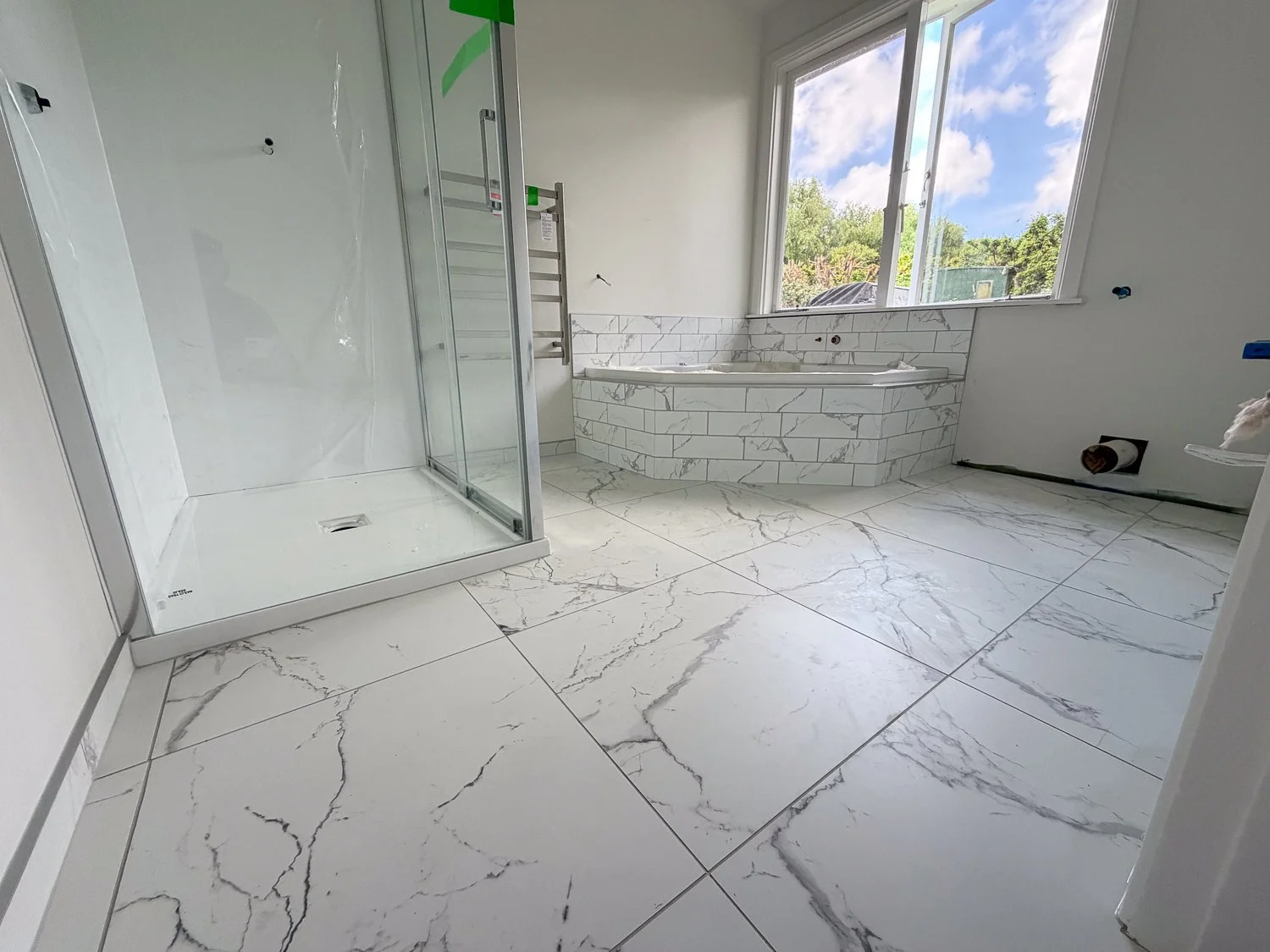 Empty modern bathroom under construction with a walk-in shower, bathtub, and large window showing outdoor scenery.