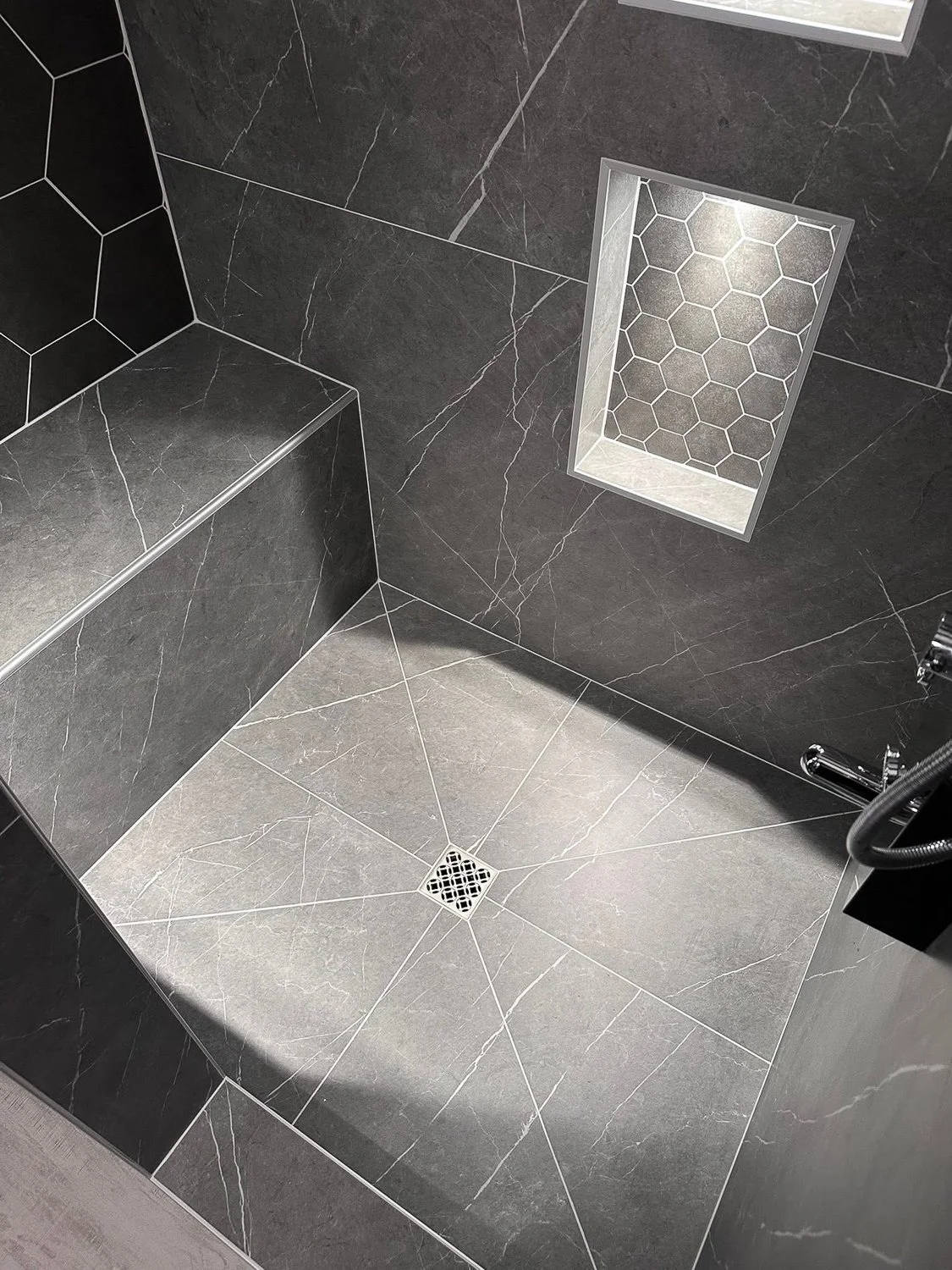 Close-up of a modern, black tiled shower with a built-in bench, a small hexagonal tile niche, and a square drain cover on the floor.
