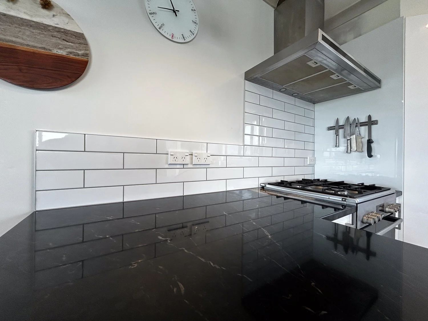 Modern kitchen with black marble countertop, white subway tile backsplash, wall-mounted knife rack, stainless steel stove, and a wall clock.