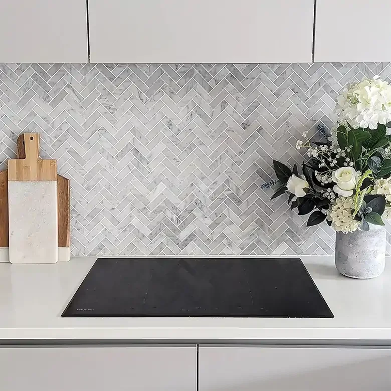 Kitchen countertop with a black stovetop, a vase of white flowers, and a wooden cutting board with a white tile background.