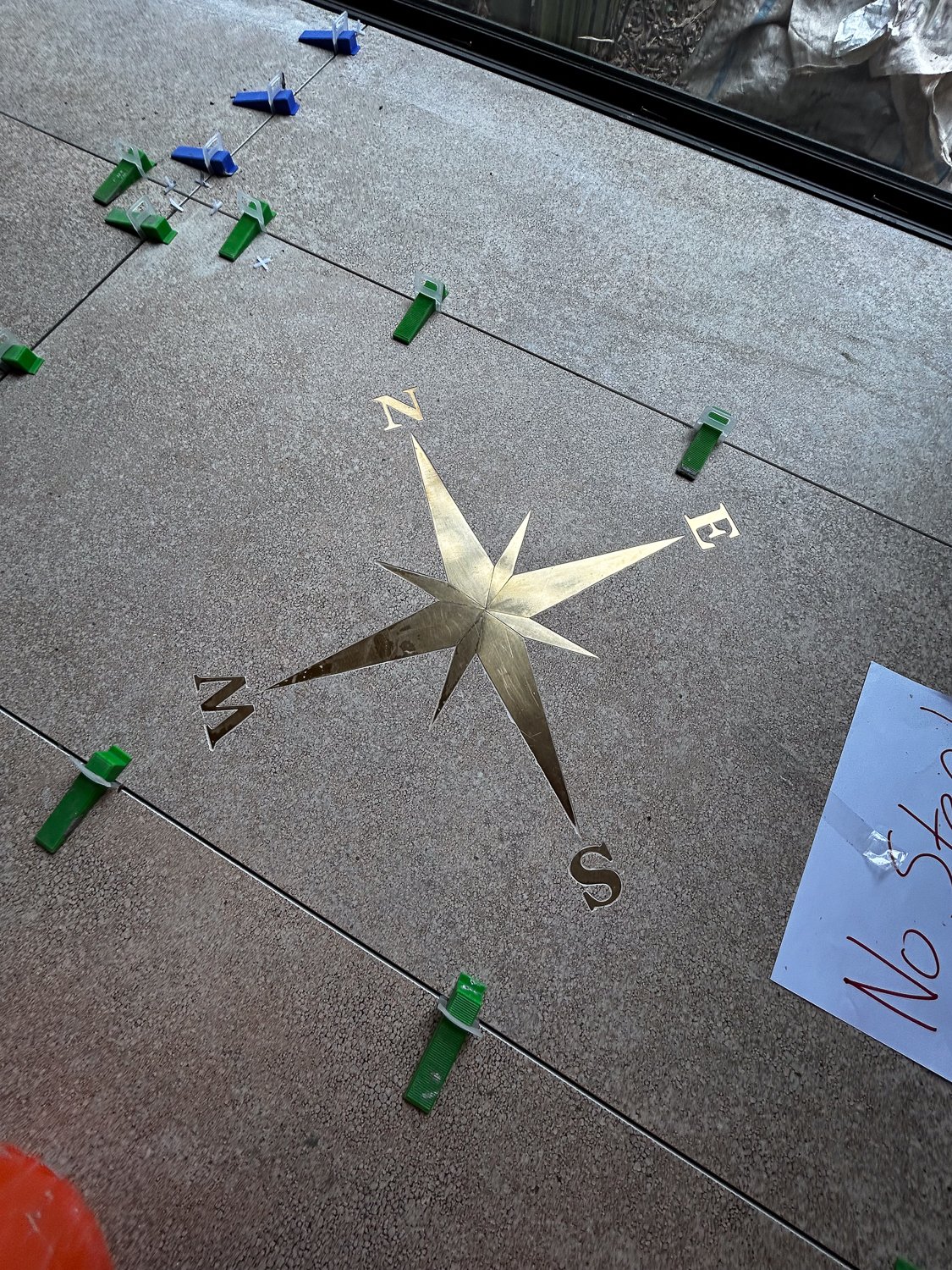 A decorative metal compass rose on a tile floor, with a gold and silver star design, surrounded by black and gold directions, and green and blue adjustable clamps on a wire line.