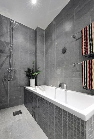 Modern bathroom with gray tiles, a white bathtub with dark gray mosaic sides, a wall-mounted shower, a potted plant, and two striped towels hanging on the wall.