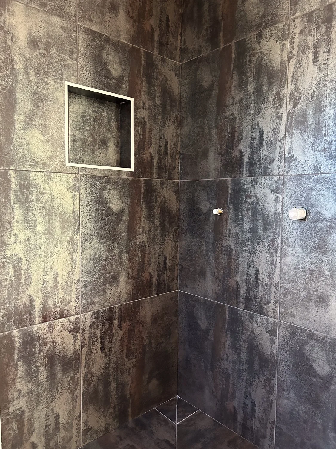 Empty shower with dark textured tiles, a recessed shelf, and plumbing fixtures for a showerhead and faucet.