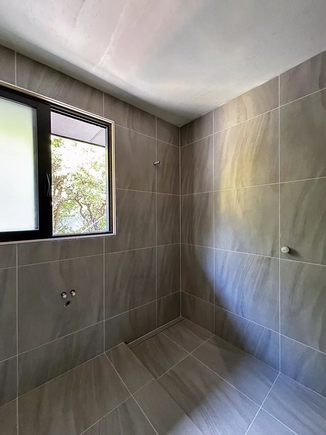 Empty bathroom with beige tiled walls and floor, window showing outdoor trees, and plumbing fixtures for a sink and shower.