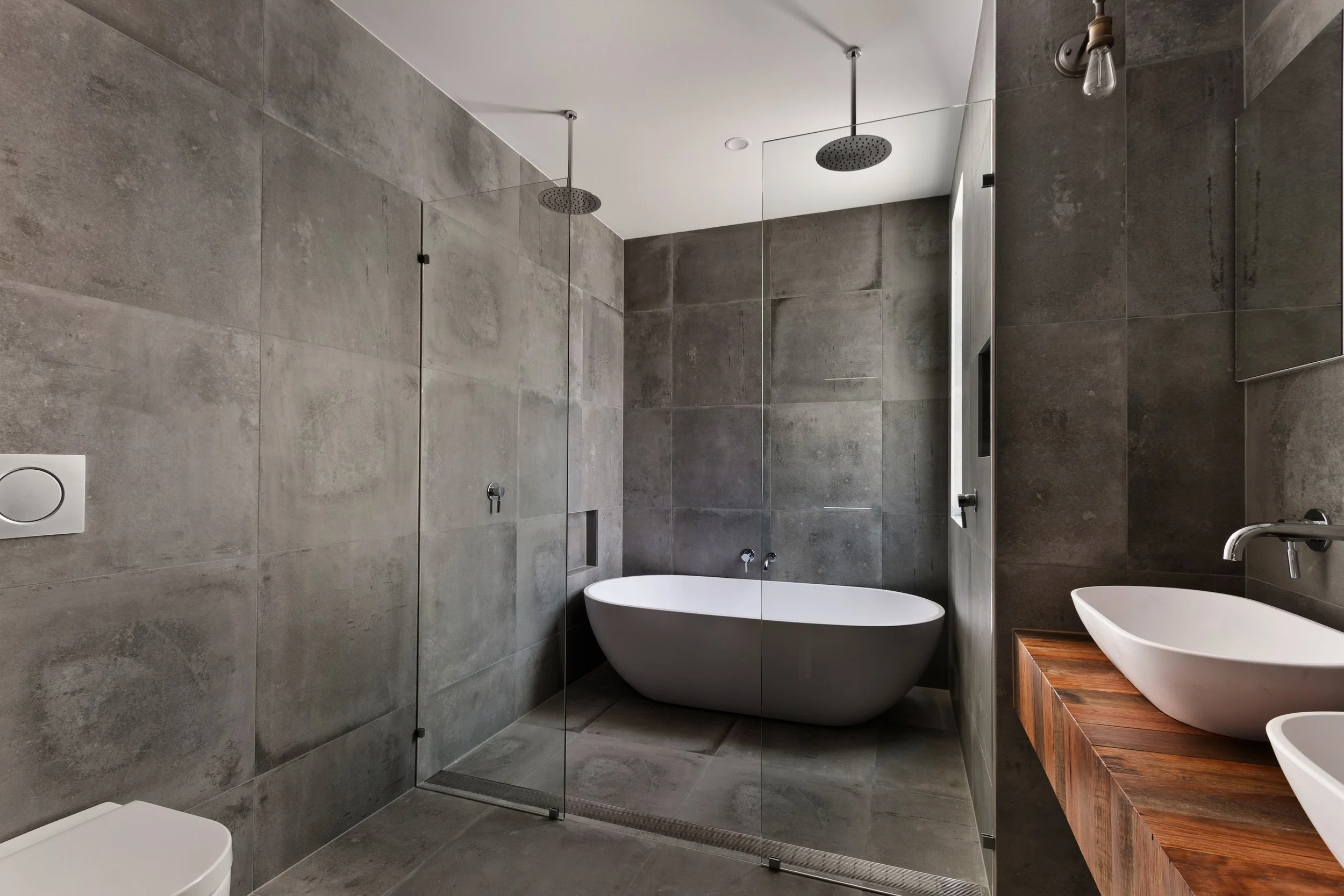 Modern bathroom with gray tiled walls, a freestanding bathtub, a glass shower enclosure with two rainfall showerheads, and a wooden vanity with two vessel sinks.