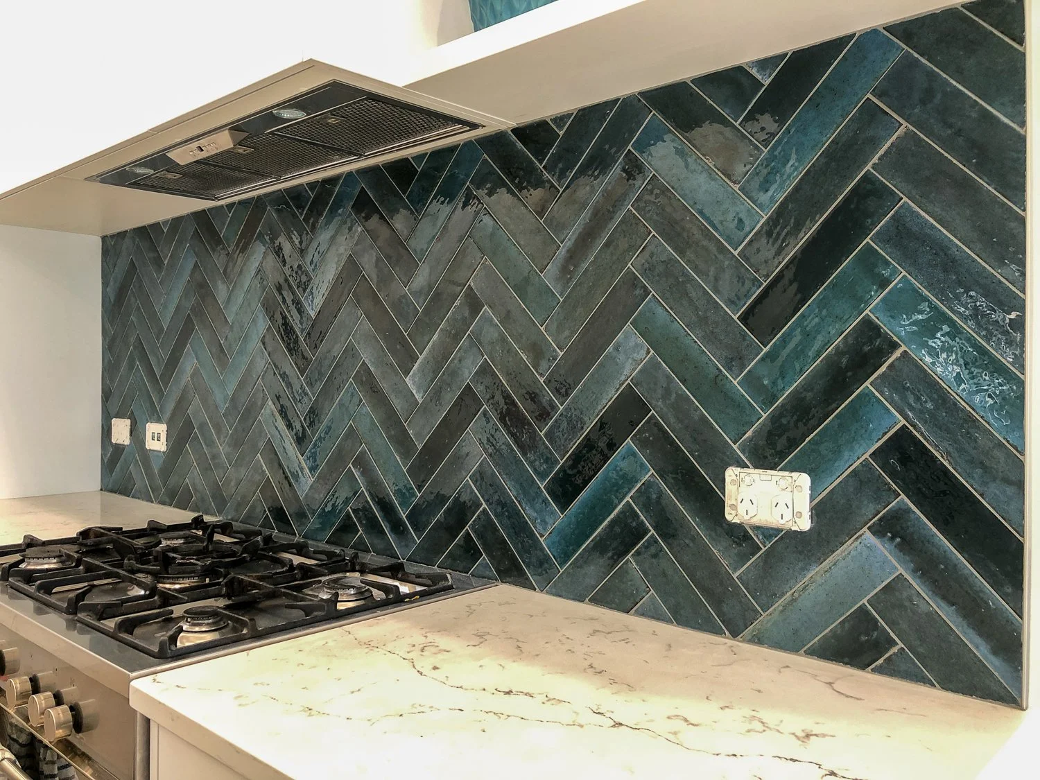 Kitchen backsplash with dark blue and teal herringbone tiles, a white marble countertop, and a stainless steel gas stove.