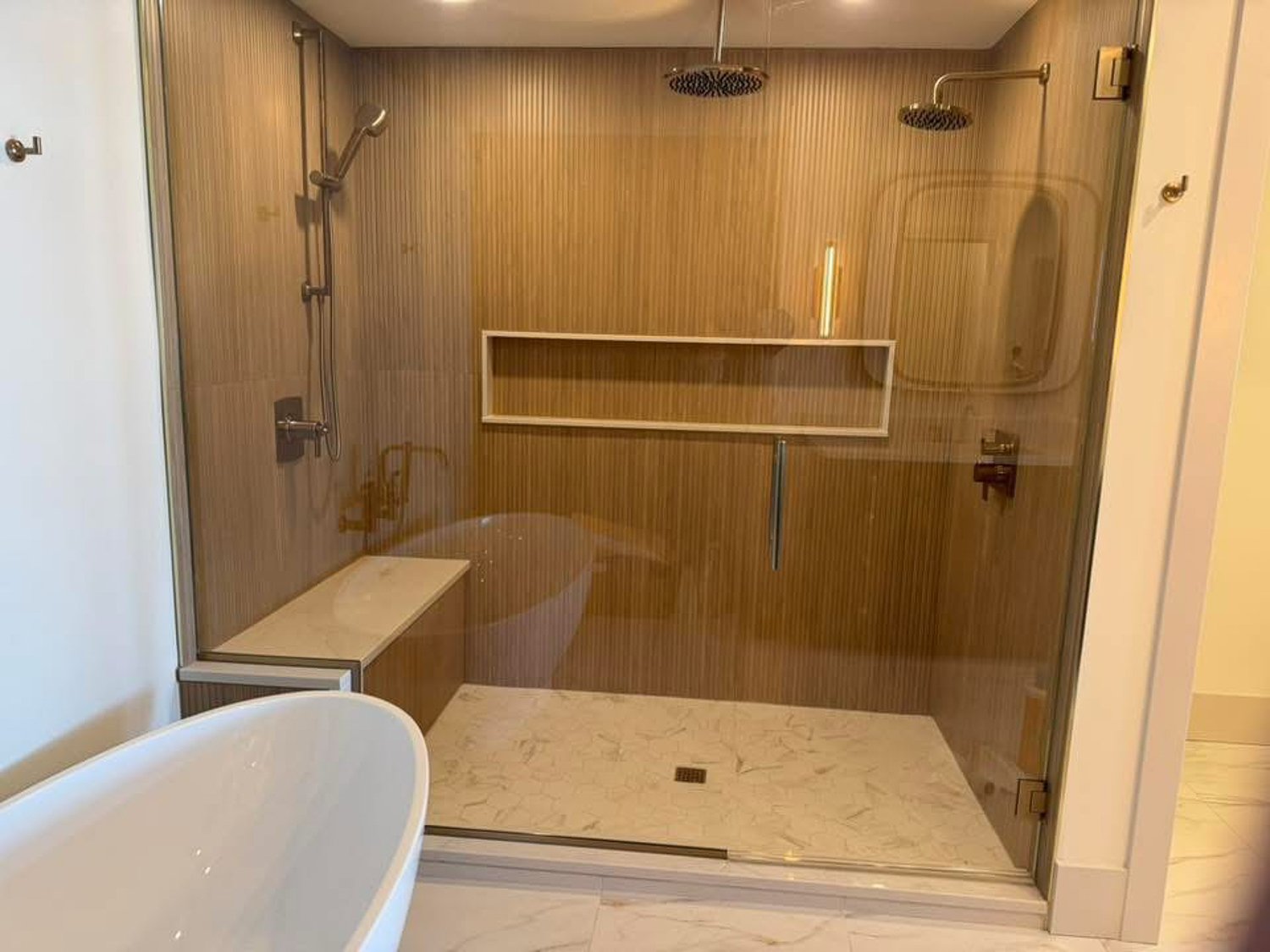 Modern shower area with wooden walls, two round rainfall showerheads, a small built-in shelf, and a marble floor drain. The shower is separated by a glass panel, and a portion of a white bathtub is visible in the foreground.