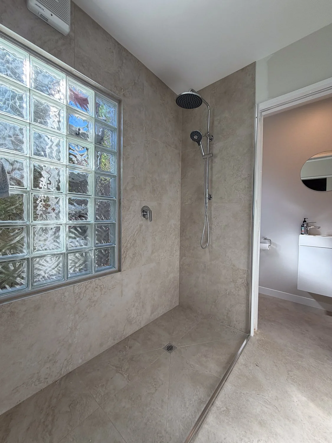 Modern walk-in shower with beige tile walls and floor, a glass block window, a ceiling-mounted showerhead, and a handheld shower wand, adjacent to a bathroom with a sink, mirror, and toilet.