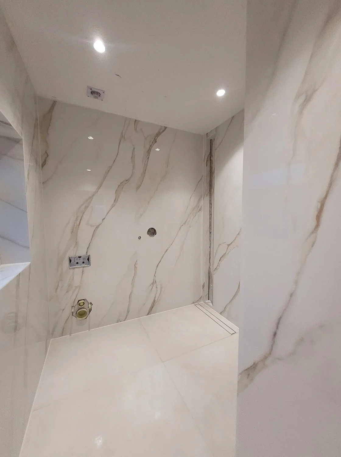 Empty shower area with marble walls, ceiling covered in white tiles with small recessed lights, and plumbing fixtures without a showerhead.