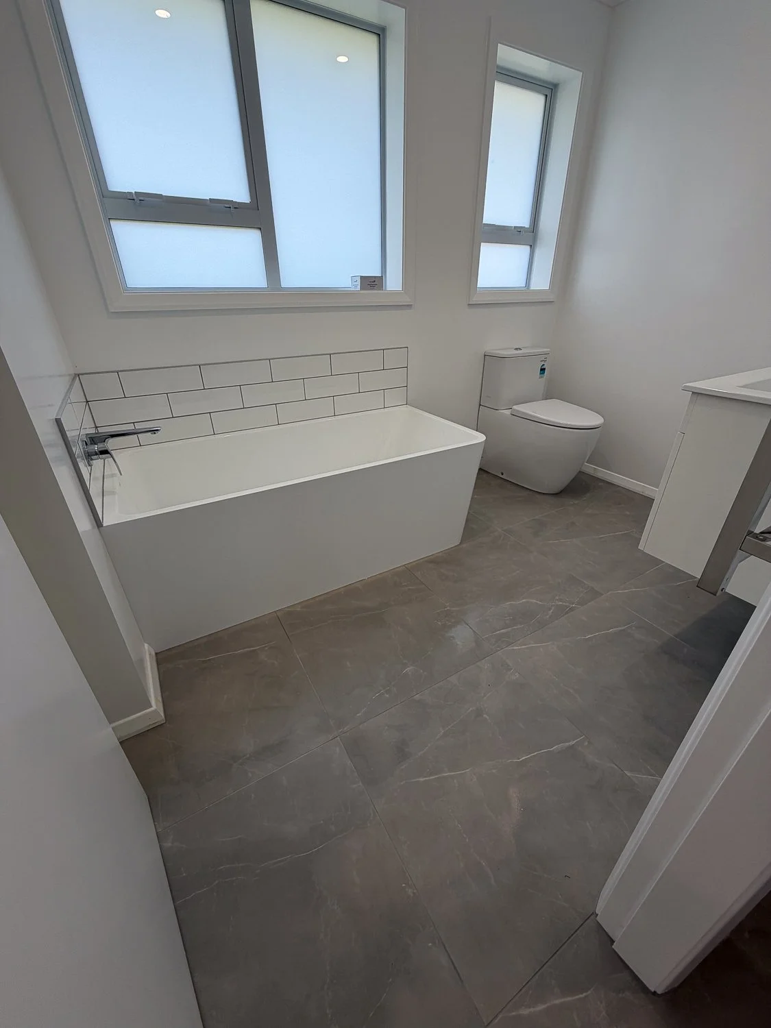 Modern bathroom with a bathtub, toilet, gray tile floor, white walls, and two frosted windows.