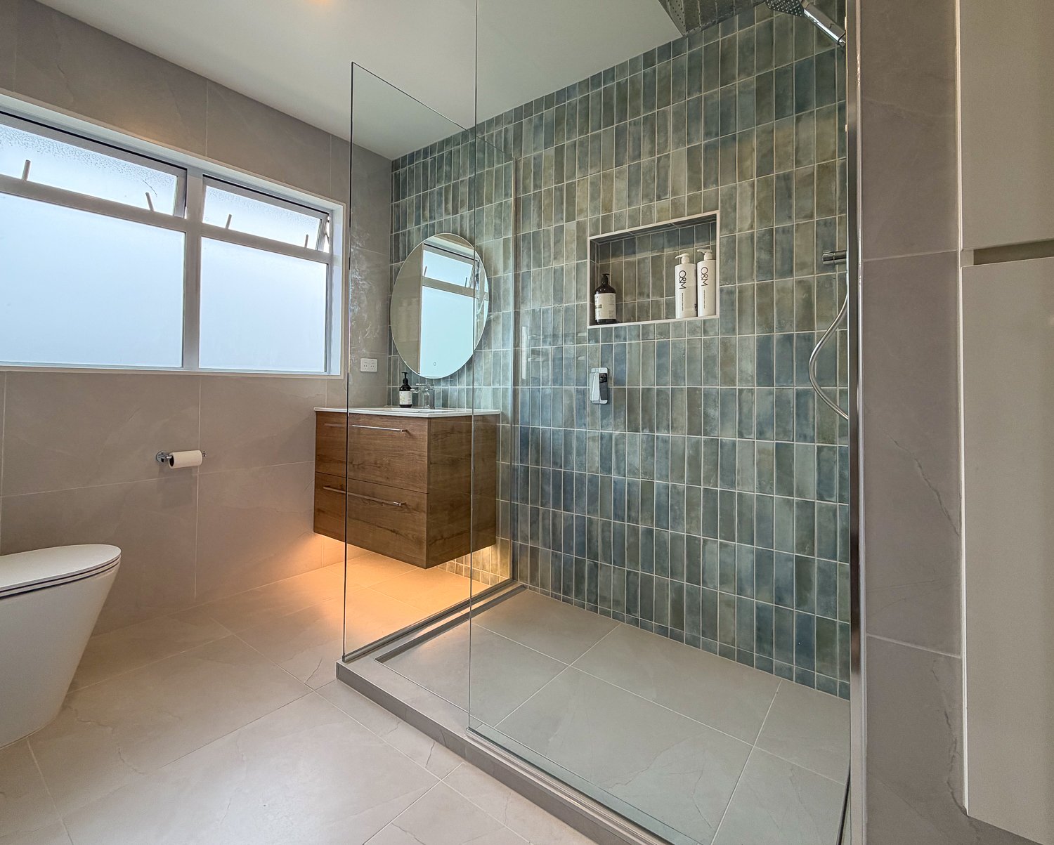 Modern bathroom with frosted window, white toilet, wooden vanity with an oval mirror, and a walk-in shower with green tiles and a glass enclosure.