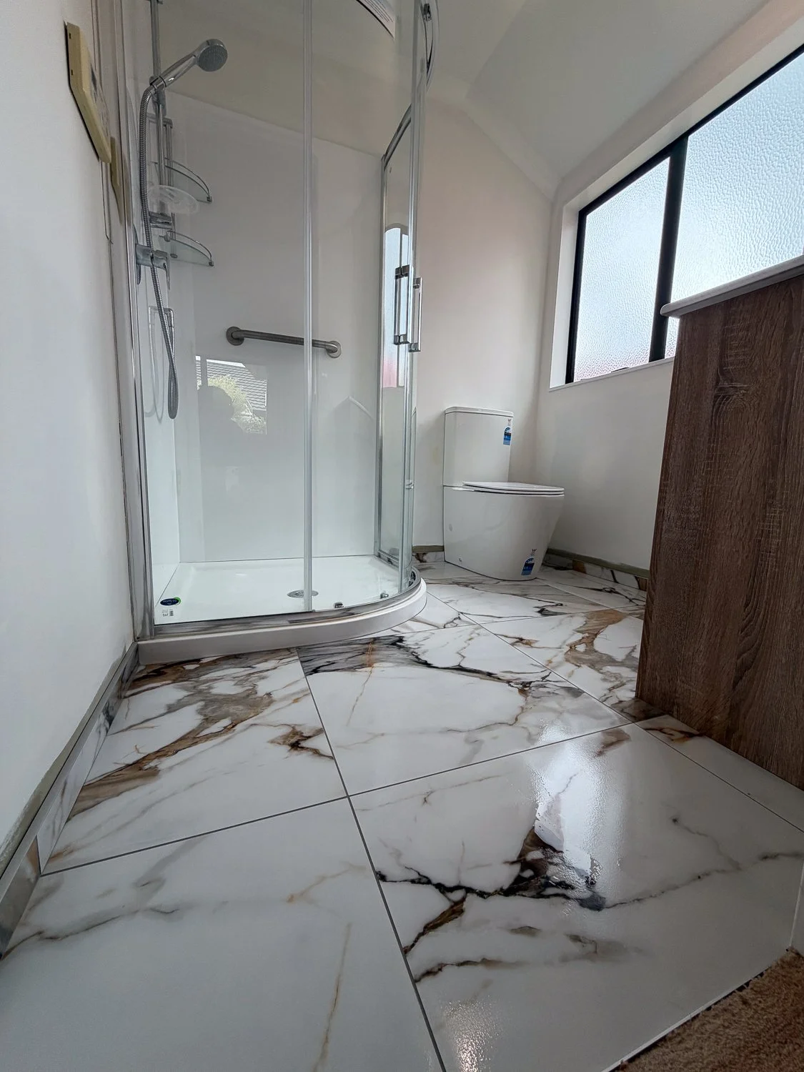 Bathroom with a glass shower enclosure, a modern toilet, and a wood cabinet. The floor is decorated with white marble tiles with brown and black veining, and a window with frosted glass lets in natural light.