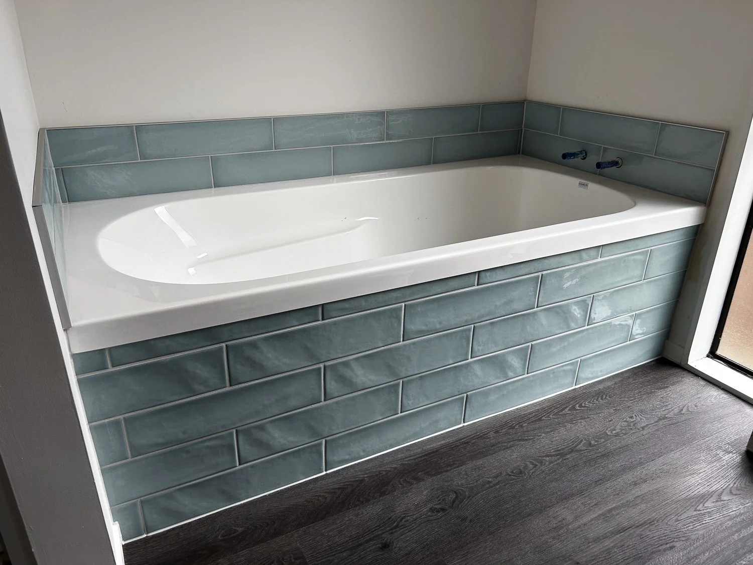 Empty bathtub with teal tiles on the surround and wall, located next to a door with a wooden floor.