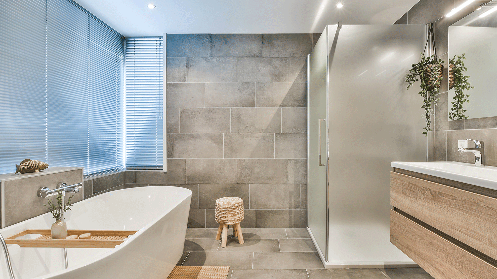 Modern bathroom with large gray tiled walls, a freestanding bathtub with a wooden tray, a stool, a vanity with a mirror and a hanging plant, and a window with blinds.