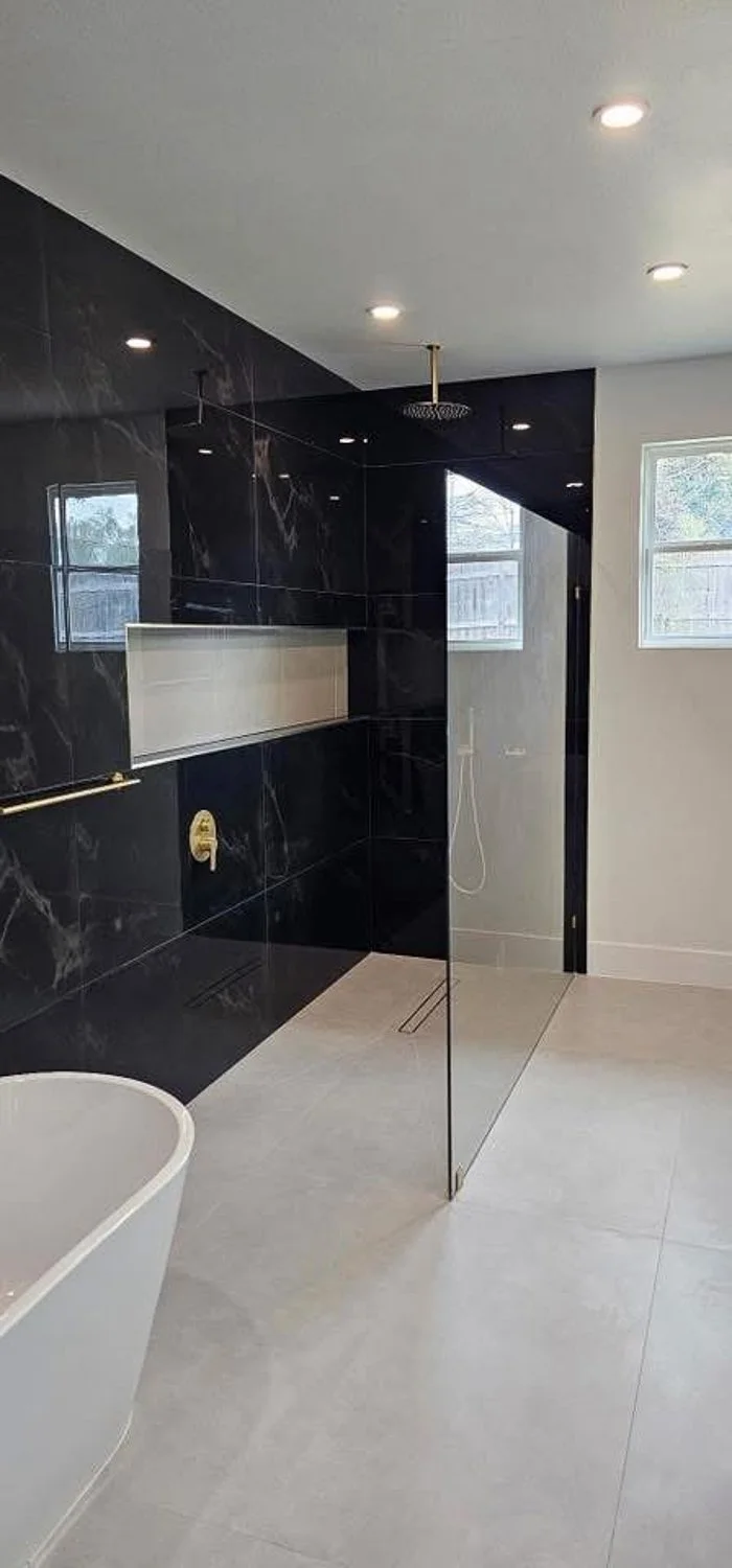Modern bathroom with black marble shower wall, glass partition, and a white bathtub, with windows providing natural light.