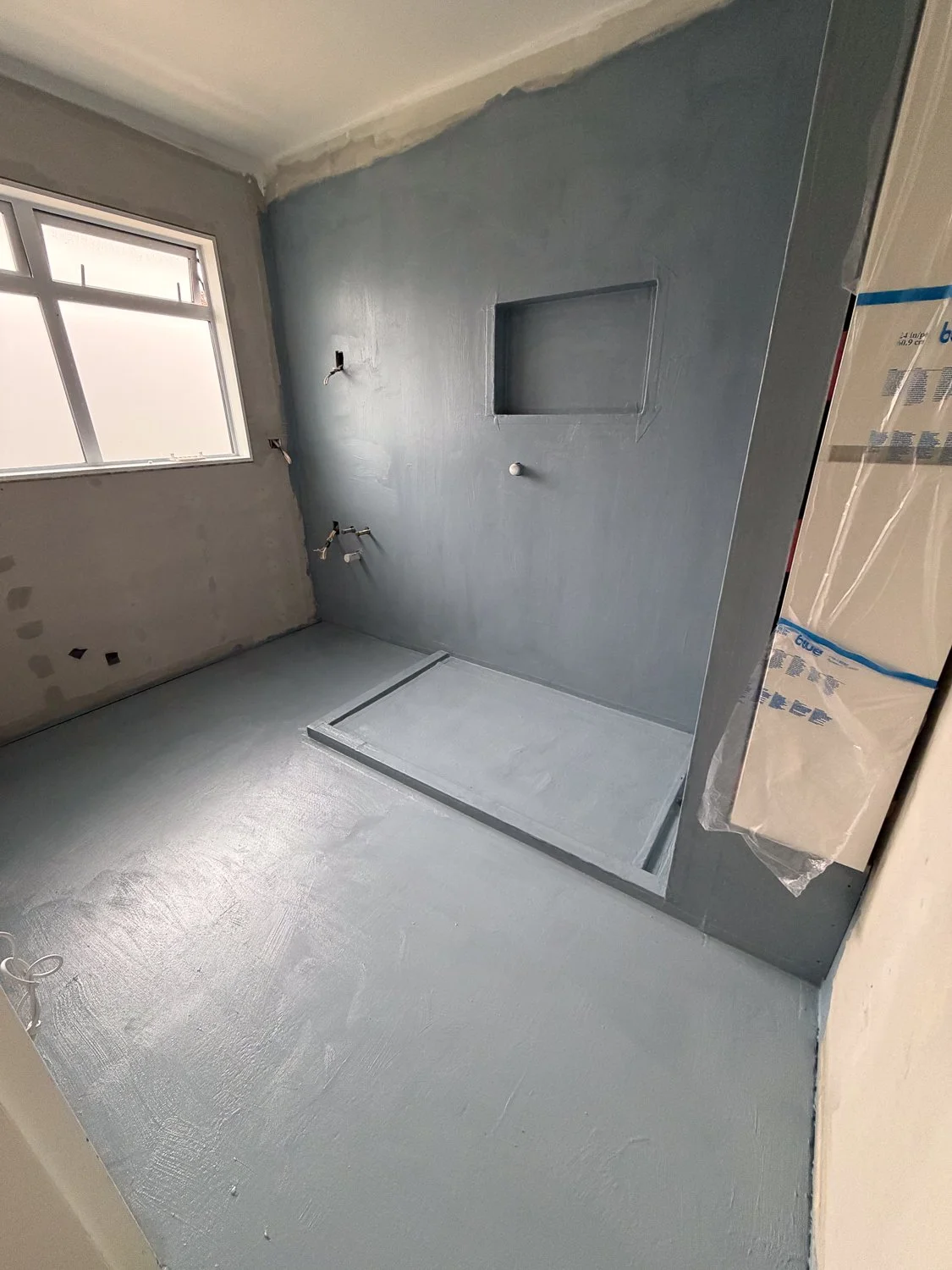 Bathroom under construction with a gray painted wall, a window, and a shower base installed, but no fixtures yet.