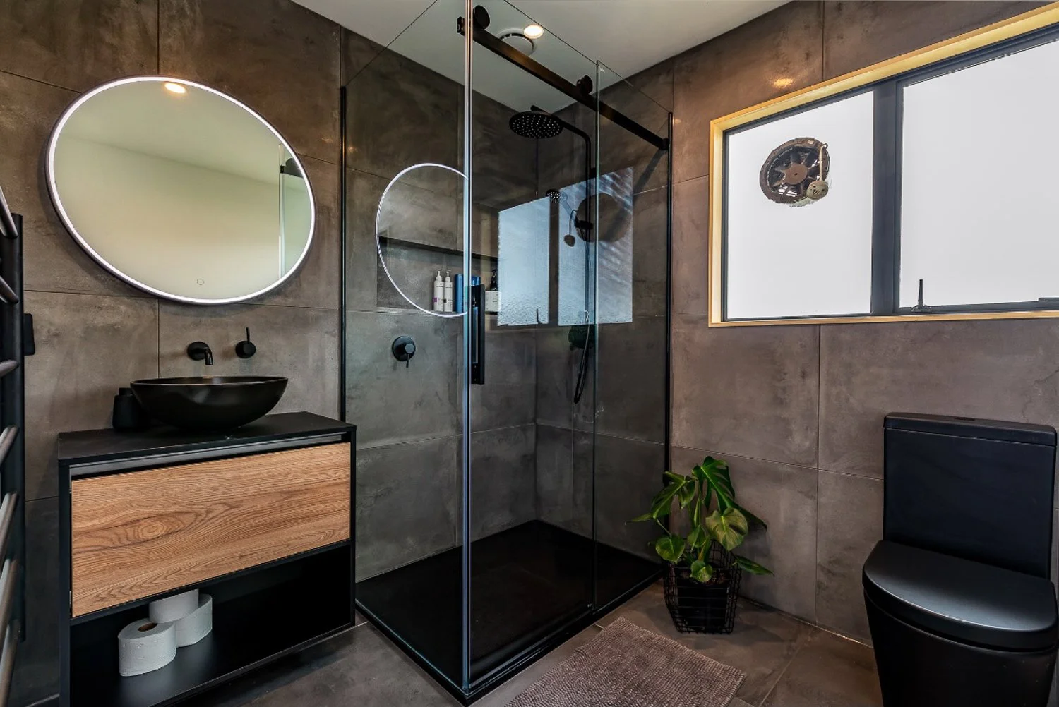 Modern bathroom with gray tiled walls and floor, a black and wood vanity with a black vessel sink, a round illuminated mirror, a glass shower enclosure with a rainfall showerhead, a window with a view outside, a black toilet, a potted plant on the fl