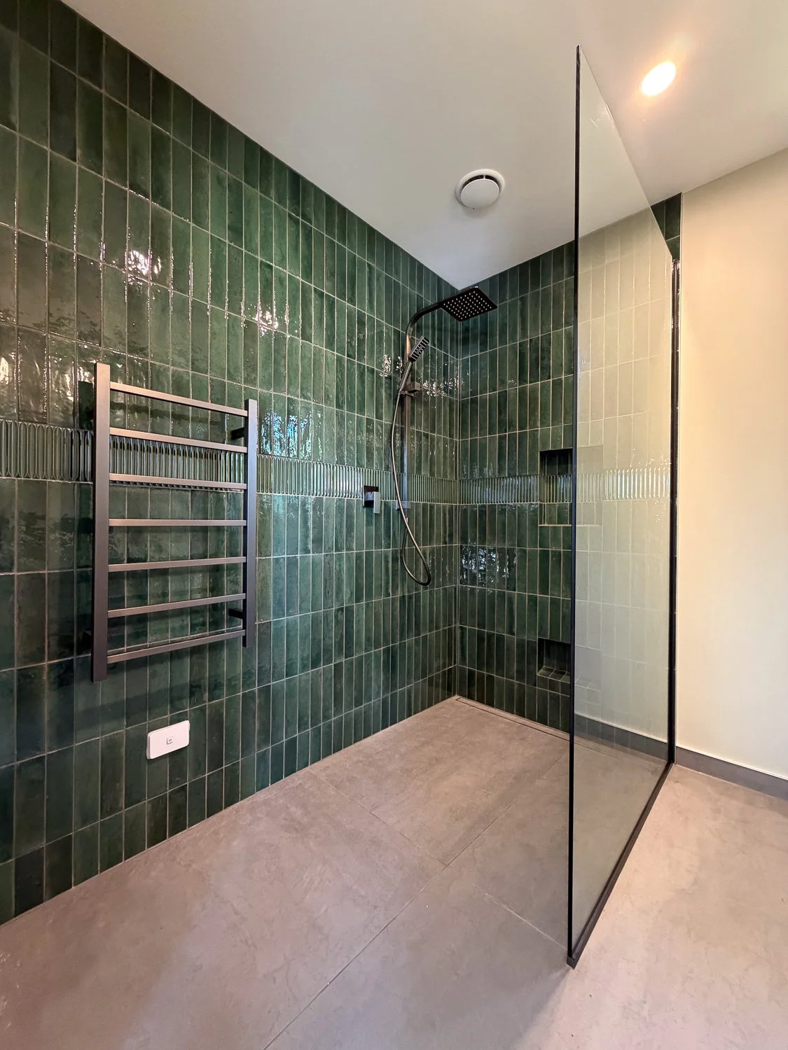 Modern walk-in shower with green tiled walls, a rainfall showerhead, a handheld shower, a niche for soap, and a metal towel rack.