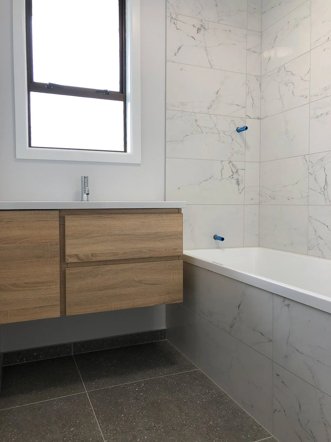 Modern bathroom with a window, wooden vanity, white sink, marble tile bathtub surround, and black tile flooring.