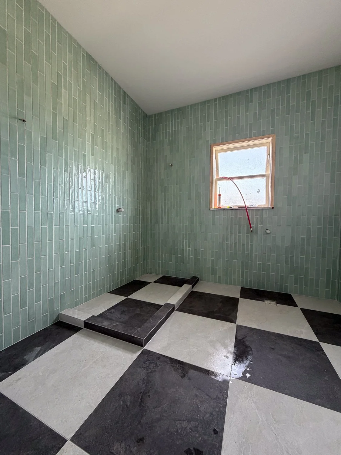 Empty bathroom with green tiled walls, black and white checkered floor, and a small window; area for a shower or bathtub under construction.