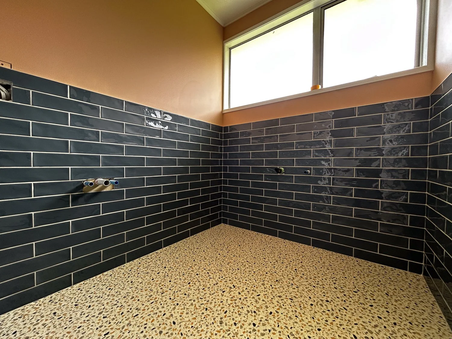 Interior of a bathroom under construction with black subway tile walls, a terrazzo floor, and a large window.