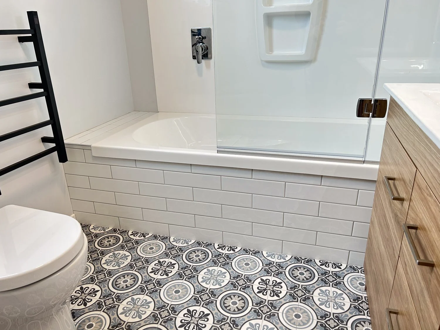 Bathroom with patterned tile floor, white subway tile wall, bathtub with glass shower door, wall-mounted faucet, wooden vanity with drawers, toilet, and black towel rack.