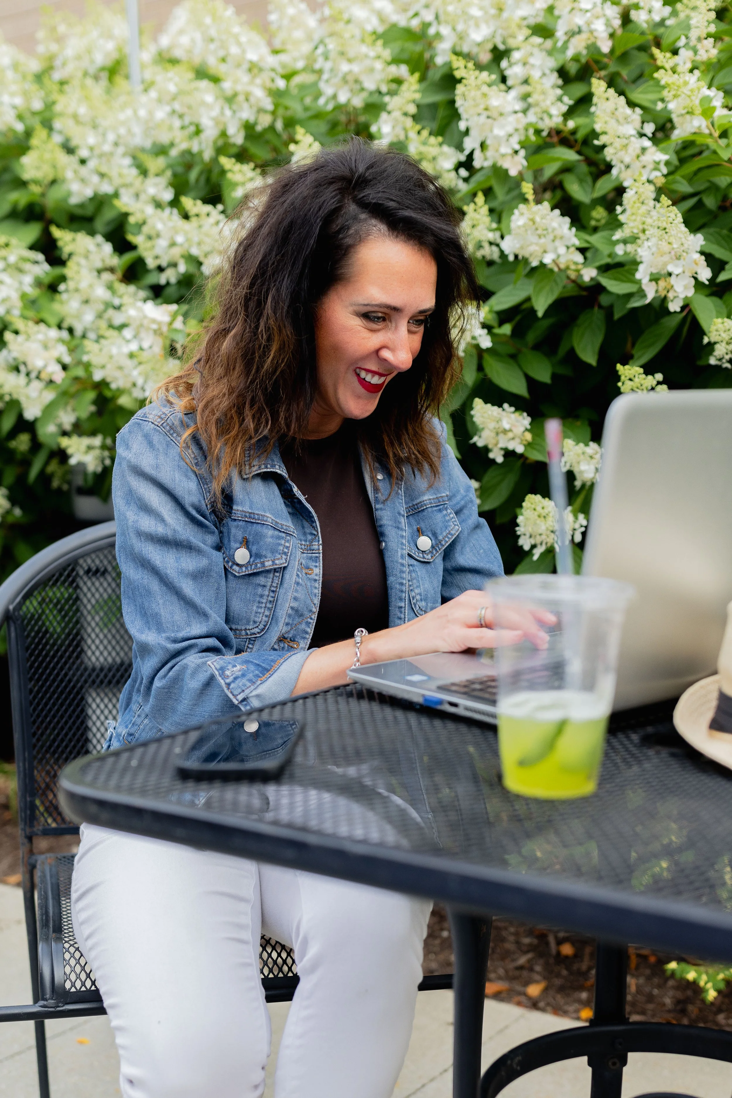 Smiling woman in a denim jacket working on a laptop at an outdoor café surrounded by white flowering shrubs
