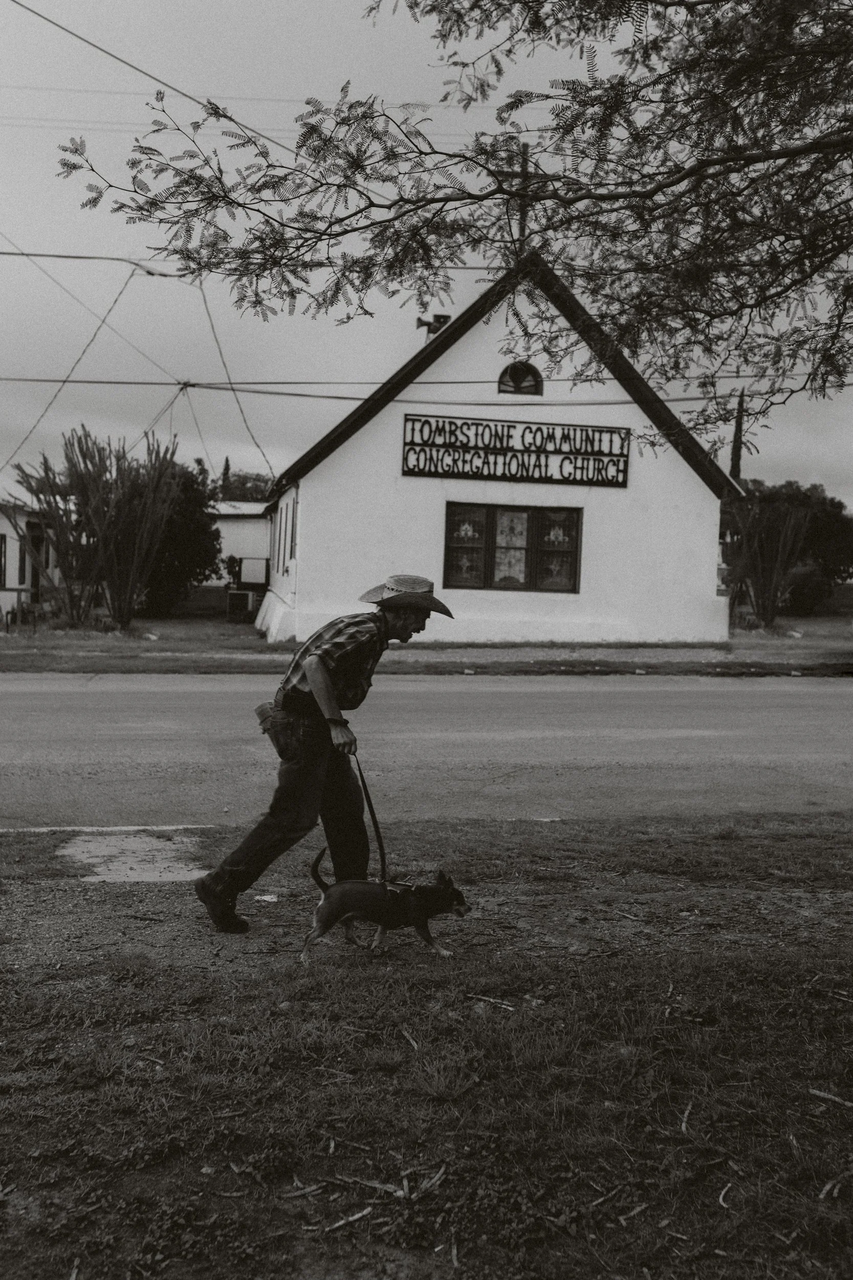 Une personne marchant avec un chien sur le trottoir devant une bâtisse blanche avec une enseigne indiquant "Tombstone Community Congregational Church", en noir et blanc.
