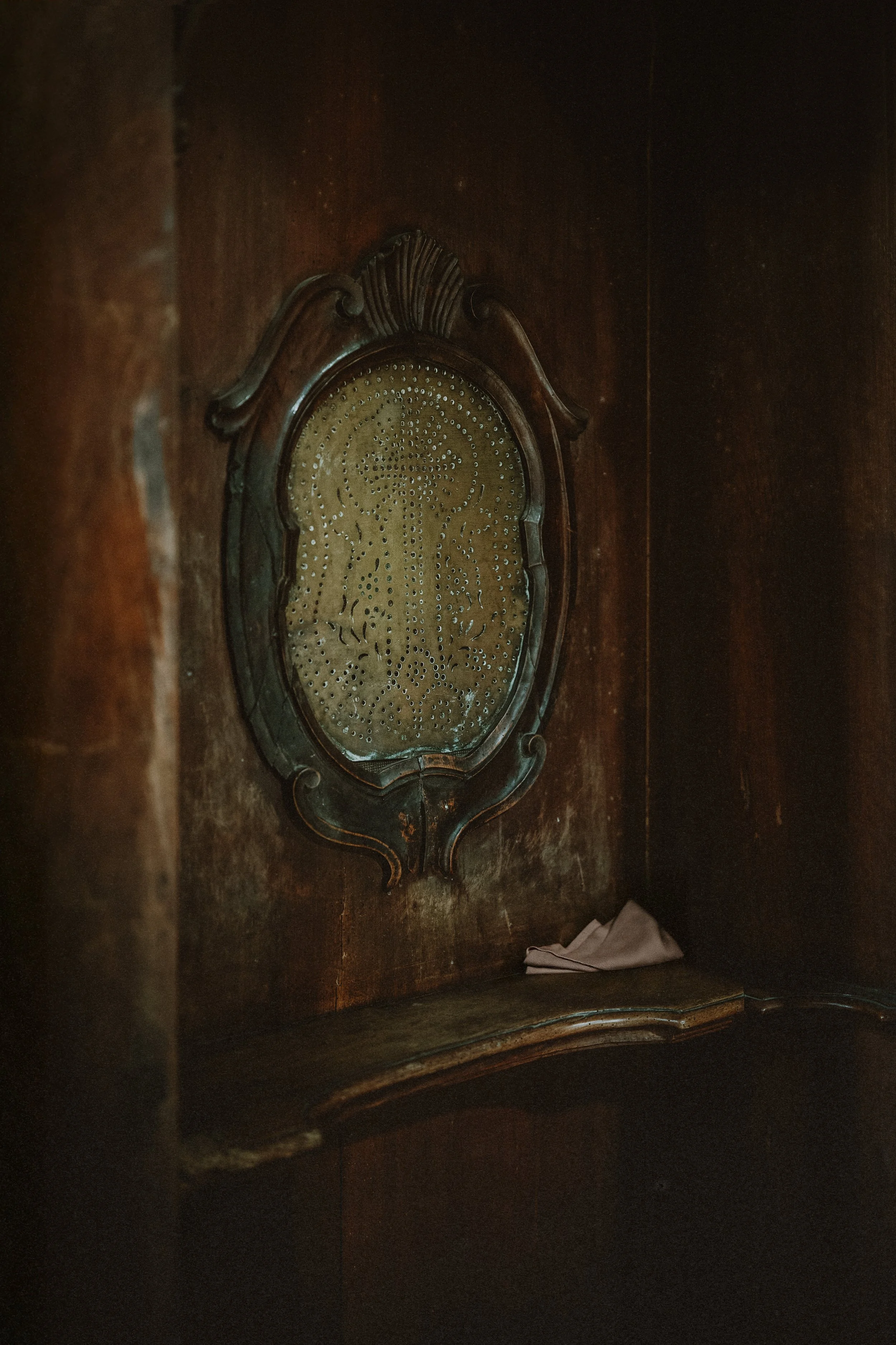 Un vieux mur en bois avec un tableau en verre orné d'une décoration en métal, une petite optique en haut, et un objet plié en tissu sur une petite étagère en bois en bas.