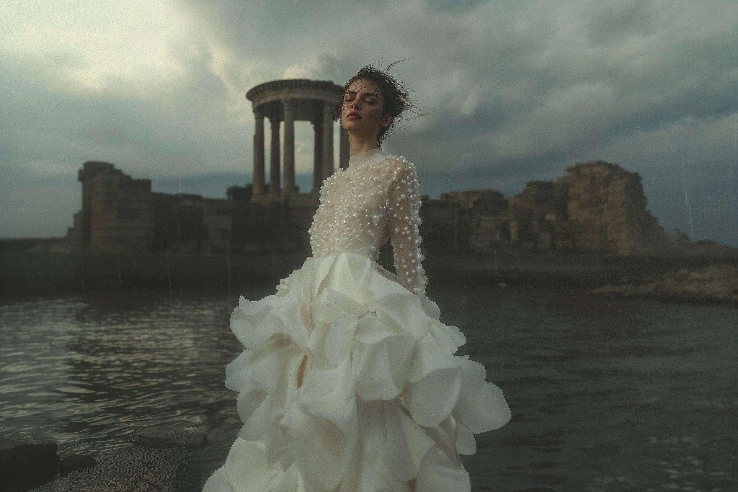 Une jeune femme portant une robe blanche ornée de perles, debout dans l'eau près d'une ruine antique sous un ciel nuageux.